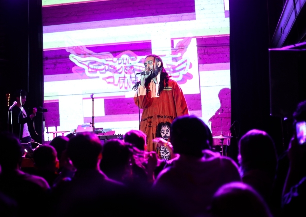 Heno performing to a large crowd with a purple striped background. 