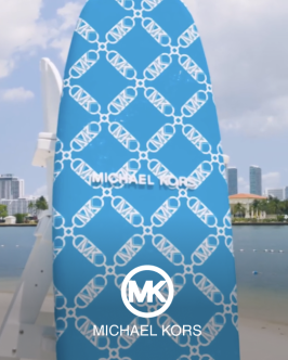 A patterned blue Michael Kors upright surfboard  in the sand on a beach with a white Michael Kors logo on top of the image
